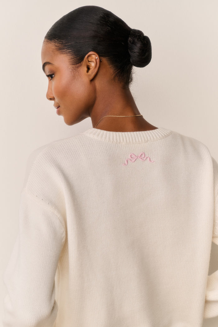 Woman wearing a cream sweater with a pink bow design on the back against a neutral background