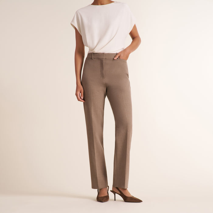 A model from the back wearing high-waisted taupe straight-leg trousers, a white draped short-sleeve top, and brown pointed-toe slingback heels against a warm off-white studio background. | Lead