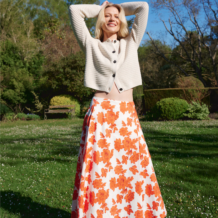 Woman in a white cardigan and orange floral skirt standing in a park. | Lead