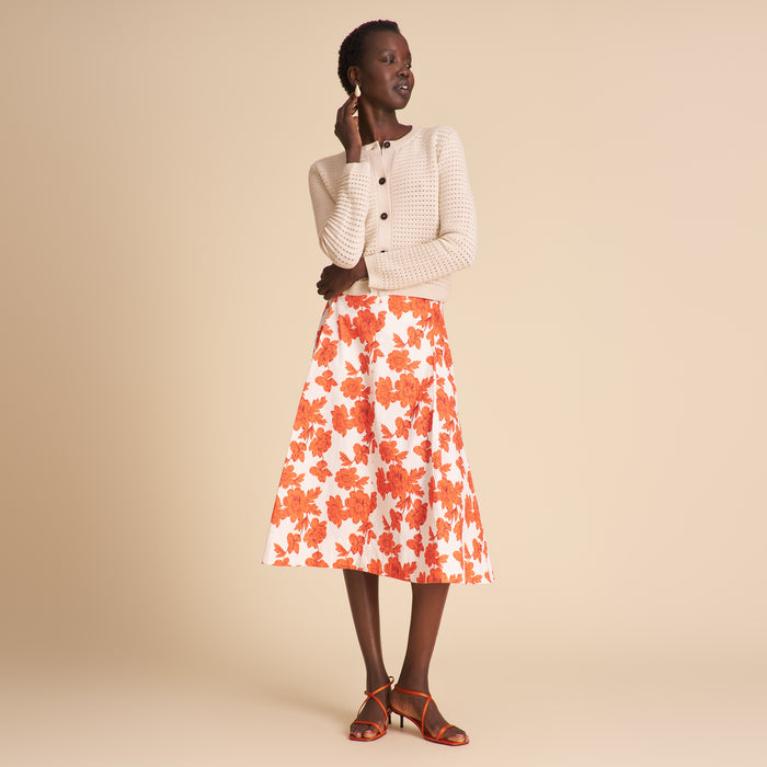 Woman wearing a white cardigan, floral skirt, and high-heeled shoes on a beige background