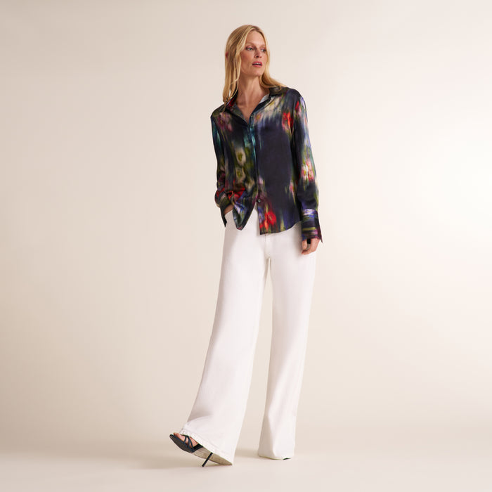 A woman modeling a long-sleeve button-up shirt with a dark abstract multicolor print, paired with white wide-leg trousers, against a plain beige studio background. | Lead