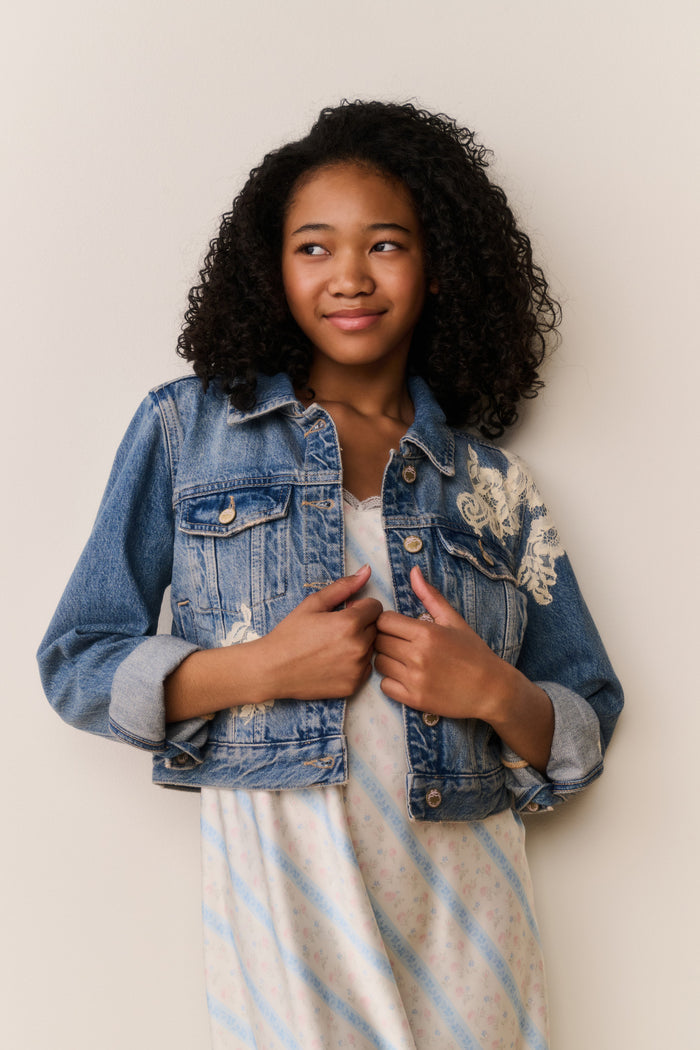 Young girl wearing a denim jacket with lace embroidery against a plain background