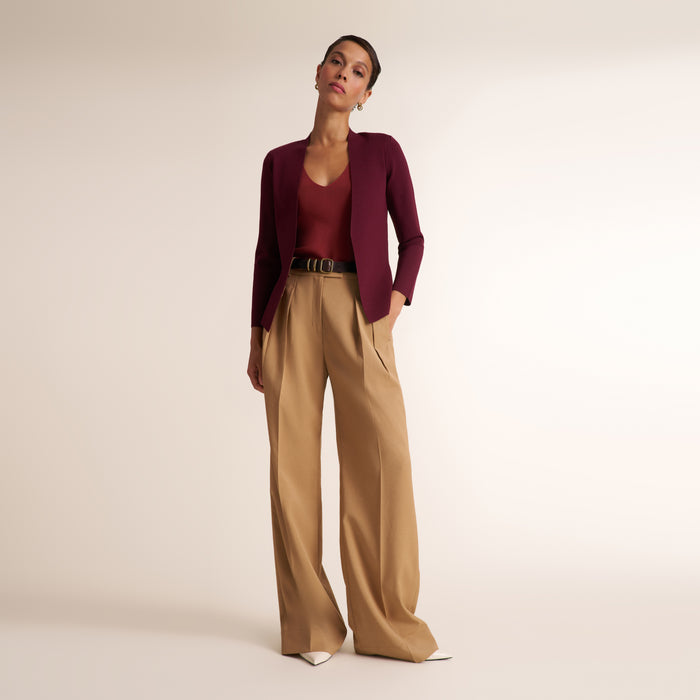A model stands against a plain beige studio background wearing a dark burgundy long-sleeve knit top, a matching dark burgundy cardigan, high-waisted wide-leg tan trousers, and white pointed shoes.