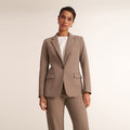A woman modeling a tailored taupe blazer with matching trousers over a white top, photographed against a plain light beige studio background. | Lead