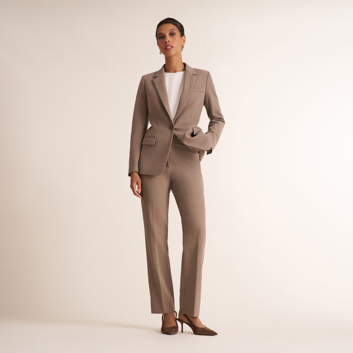 A model wearing a tailored taupe-brown two-piece suit with a blazer and matching trousers, styled over a white top and brown heels, standing against a plain light beige studio background.