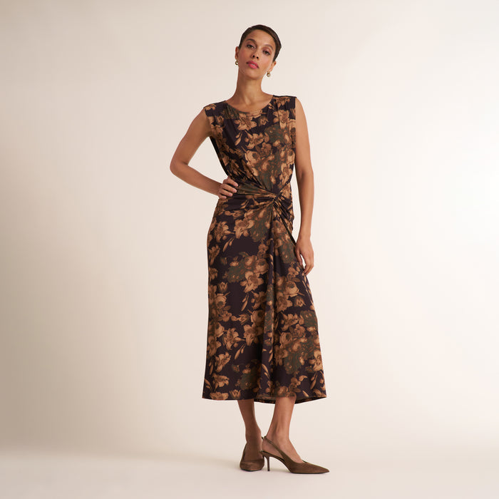 A woman models a sleeveless midi dress with a dark brown and black floral pattern and a twisted waist detail, paired with brown pointed-toe heels against a plain beige studio background. | Lead