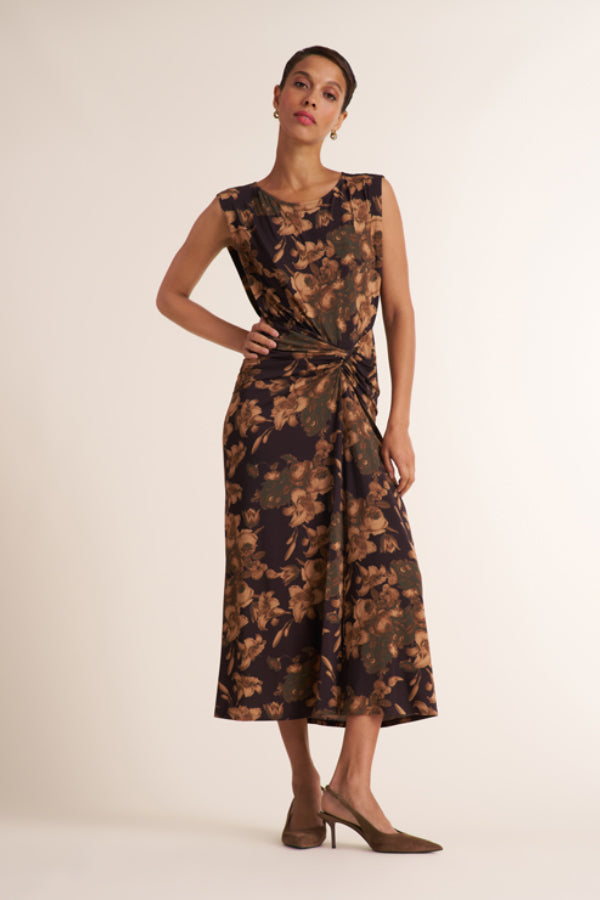 A woman models a sleeveless midi dress with a dark brown and black floral pattern and a twisted waist detail, paired with brown pointed-toe heels against a plain cream studio background. | Exclude | Grid